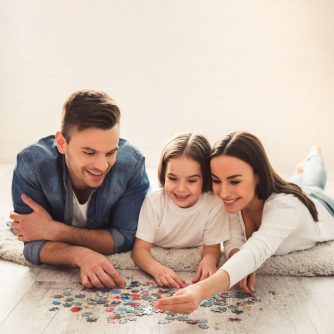 Charming little girl and her beautiful young parents are doing jigsaw puzzle and smiling while lying on the floor at home